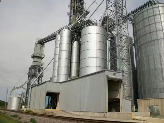 Large industrial grain elevator and feed mill facility with multiple metal silos, conveyors, and support structures next to railroad tracks.