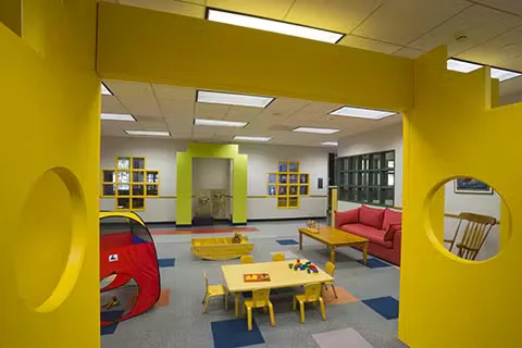 Brightly painted daycare playroom with yellow walls, child-sized tables and chairs, colorful toys, and a small play tent in a safe, open indoor space.