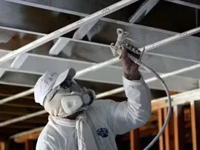 Industrial painter in full protective gear spray-painting an exposed ceiling structure with a professional spray gun inside a commercial building.