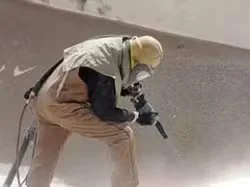 Commercial painter wearing protective gear sandblasting or surface-prepping a wall indoors, creating dust as part of an industrial painting process.
