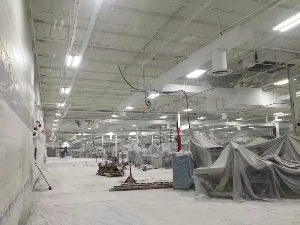 Large manufacturing facility interior with industrial equipment covered in plastic while ceilings and structural elements are professionally spray-painted during a commercial renovation.