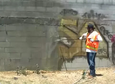 Worker wearing a safety vest uses a pressure washer to remove graffiti and grime from a concrete block wall as part of a commercial cleaning project.