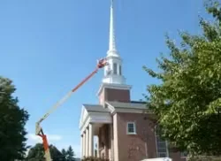 Lift equipment positioned beside a historic church steeple as workers perform exterior painting and restoration on a landmark building.