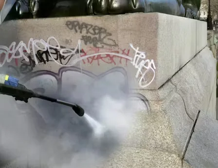 Worker pressure washing graffiti from a stone monument base as anti-graffiti coating is applied or restored, with water spray removing paint from the surface.