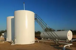Large white industrial storage tanks with metal access stairs and piping under a clear blue sky.