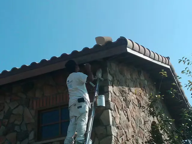 Painter standing on a ladder applying paint to the eaves of a stone building under a clear blue sky.