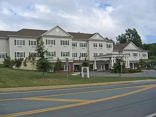 Exterior of a multi-story assisted living facility with light-colored siding, landscaped grounds, and a covered main entrance along a roadway.