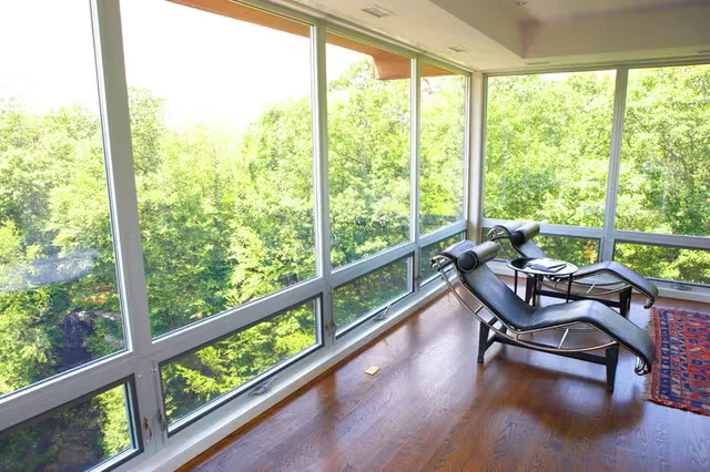 Sunlit room with floor-to-ceiling aluminum window frames overlooking green trees, featuring modern lounge chairs on hardwood flooring.