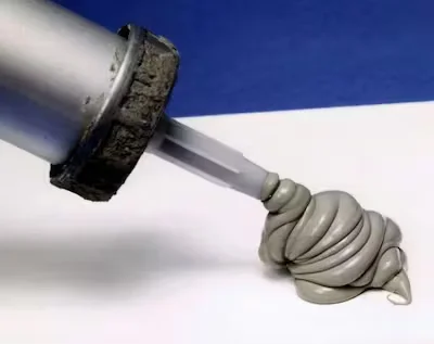 Gray construction sealant being dispensed from a caulking gun onto a white surface, forming a coiled bead.
