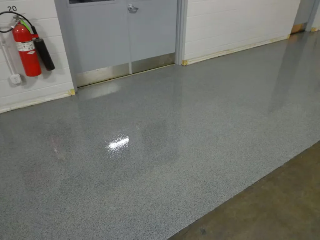 Gray epoxy floor coating in an industrial hallway with a smooth, glossy finish, adjacent to a metal door and white cinderblock walls.