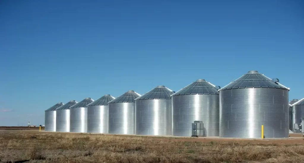 Row of large galvanized steel grain silos standing in an open field under a clear blue sky.