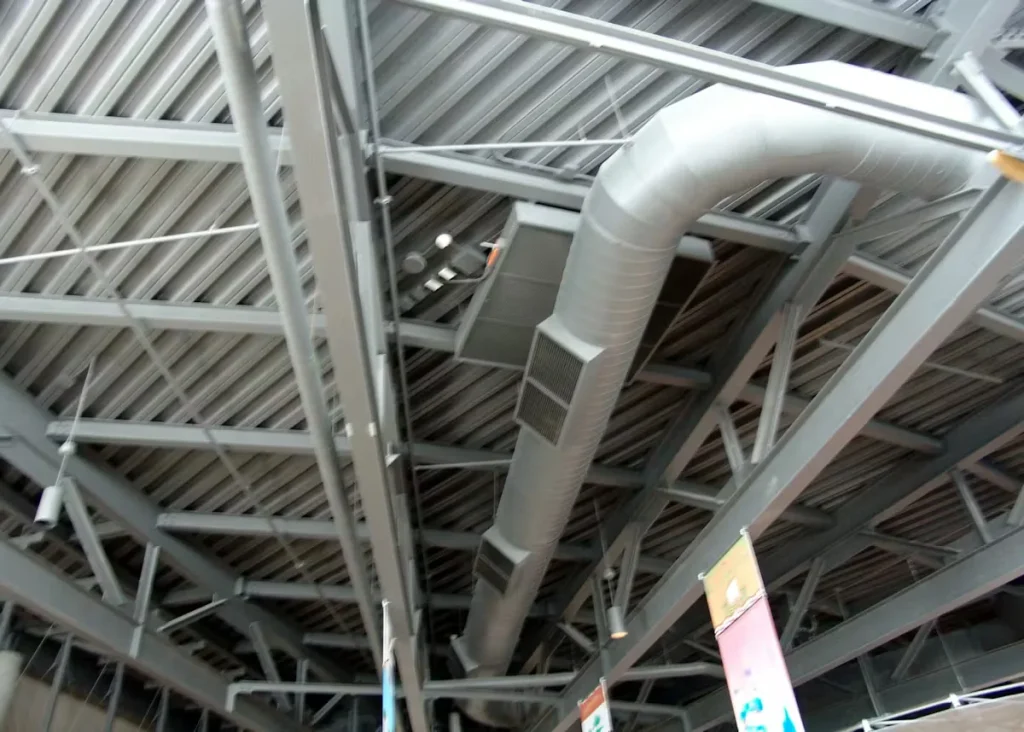 Industrial facility ceiling with exposed steel beams, corrugated metal decking, and large HVAC ductwork painted in light gray.