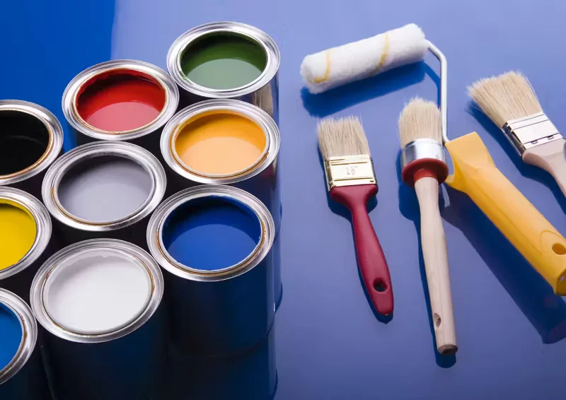 Open paint cans in various colors arranged beside paintbrushes and a paint roller on a blue surface, representing interior and exterior painting tools and materials.