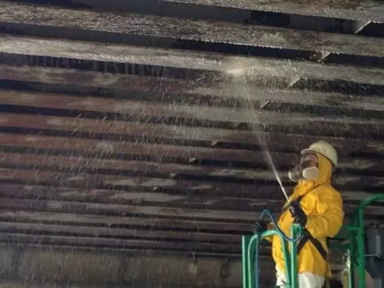 Worker in protective gear pressure-washing and degreasing the underside of an industrial ceiling or structure, removing heavy grime during industrial cleaning services.