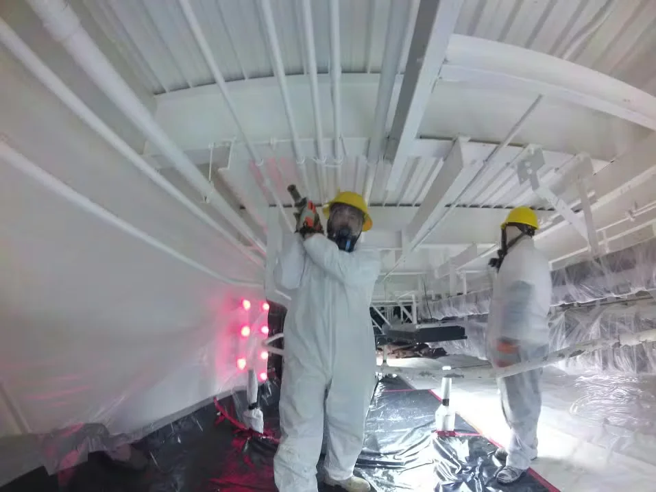 Workers in protective suits and respirators spray-painting the ceiling of a food processing facility, with exposed pipes, plastic sheeting, and safety lighting visible.