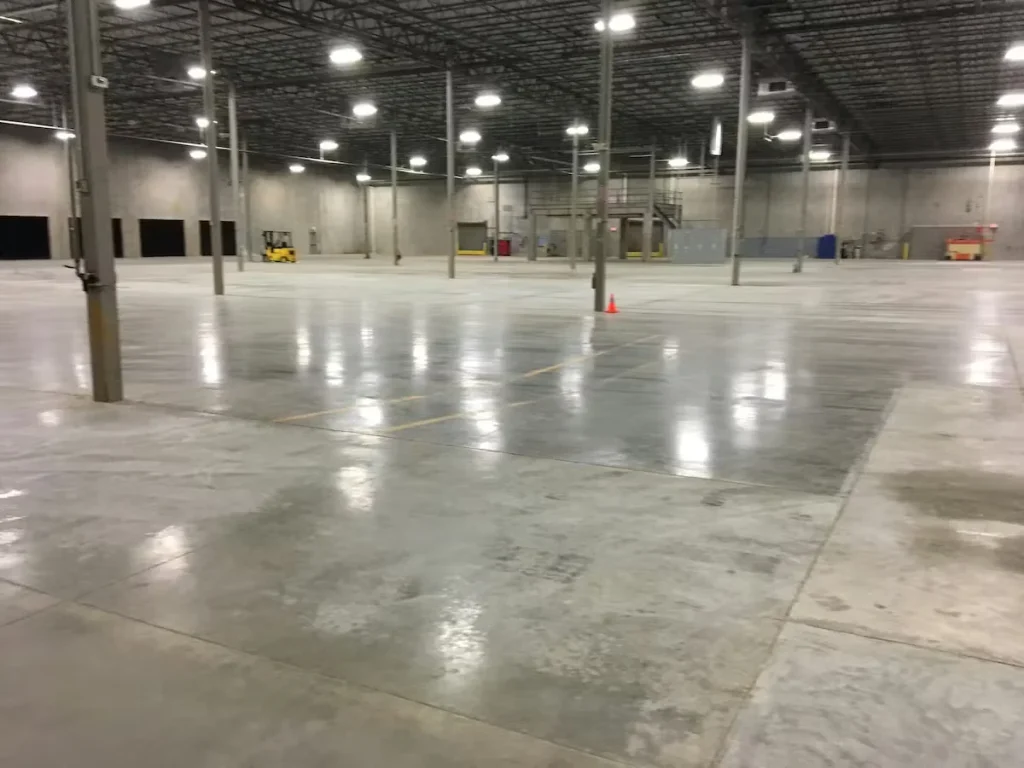 Large industrial warehouse with polished concrete floors, tall support columns, and bright overhead lighting creating reflective surfaces across the open space.