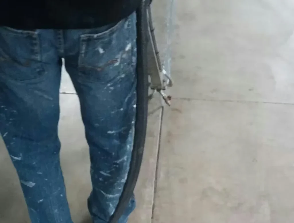 Worker wearing paint-splattered jeans standing next to a concrete floor expansion joint with exposed metal and sealant during repair work.