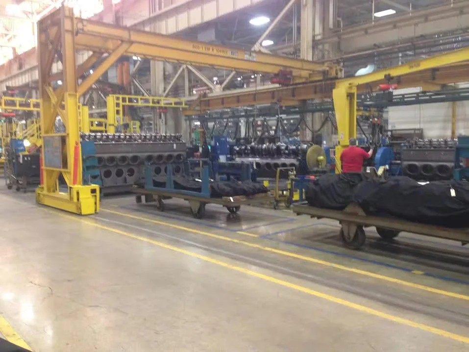 Interior of a manufacturing facility with large industrial machinery, overhead cranes, and workers operating equipment along marked production lanes on a concrete factory floor.
