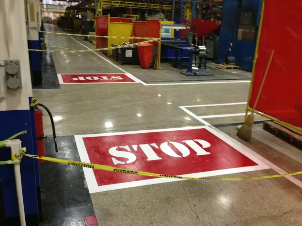 Industrial facility floor with freshly painted red “STOP” safety markings, white lane striping, and caution tape blocking access during floor painting and safety line installation.