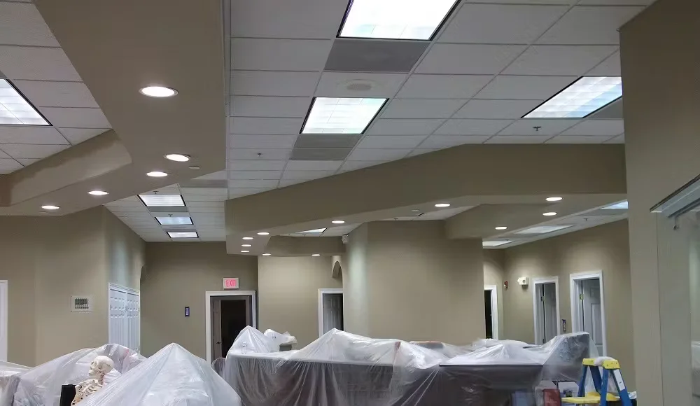 Commercial interior space under renovation with drop ceiling tiles, recessed lighting, and furniture covered in protective plastic during low- or zero-VOC painting.