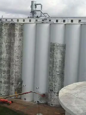 Industrial storage silos being repainted, with a boom lift positioned at the base and weathered concrete surfaces visible alongside freshly coated metal tanks.