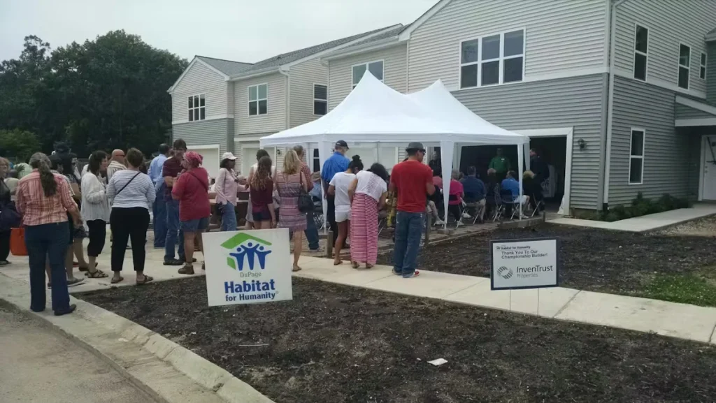 Community gathering outside a newly built duplex during a Habitat for Humanity event, with attendees standing under a white tent and DuPage Habitat for Humanity signage visible.
