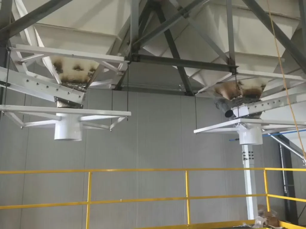Overhead equipment and steel framing inside an agricultural or food processing facility, showing dust accumulation around hoppers and duct connections above a safety railing.