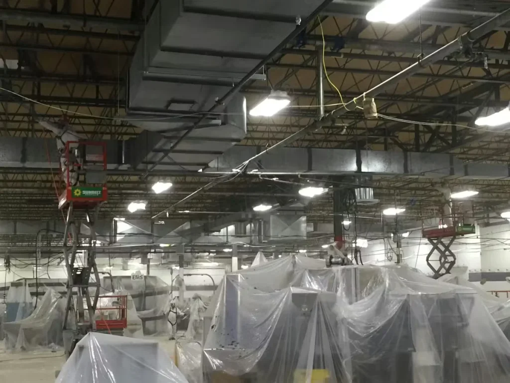 Industrial facility interior during a shutdown, showing overhead ductwork and lighting, plastic-covered machinery, and workers on lifts performing cleaning and maintenance.