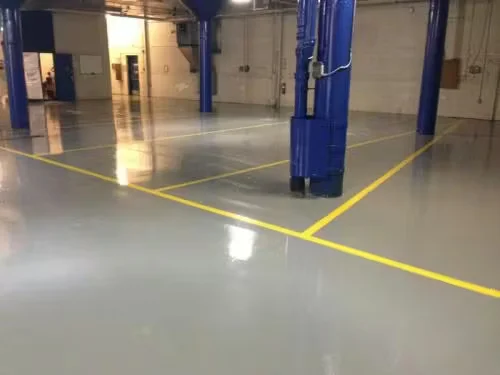 Industrial facility interior with a smooth gray epoxy floor, bright yellow safety lines, and blue support columns, showcasing a freshly coated industrial flooring system.