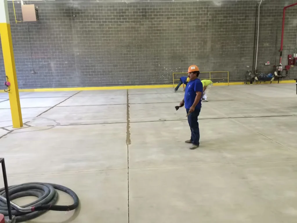 Workers applying epoxy flooring and repairing expansion joints in a warehouse storage area.