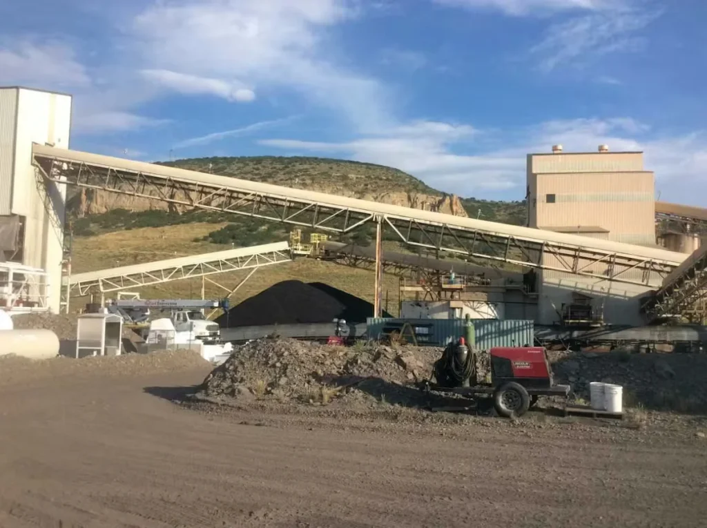 Industrial mining facility with conveyor systems, processing buildings, and equipment set against a rocky hillside.