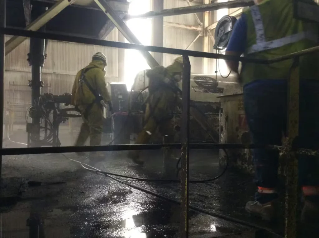 Industrial cleaning crew wearing protective gear performing high-pressure cleaning inside a manufacturing facility during equipment maintenance.