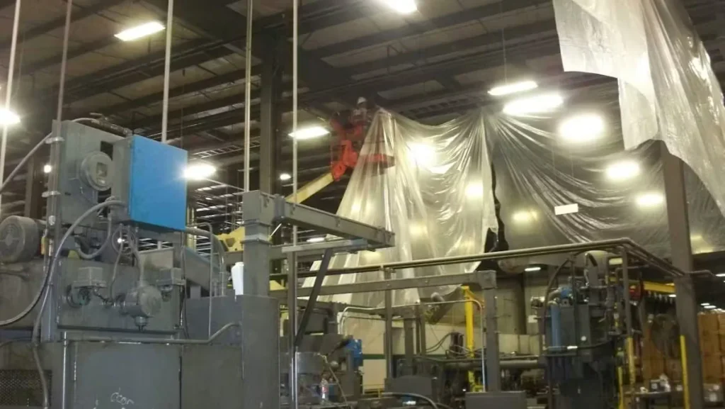 Industrial cleaning crew working inside a manufacturing facility, using lifts and protective plastic sheeting to contain debris while cleaning heavy machinery and overhead structures.