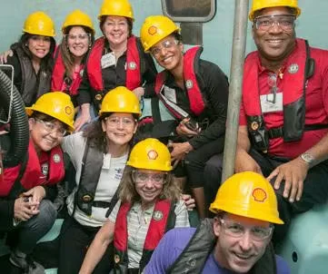 Group of workers wearing yellow hard hats and red safety vests smiling together inside an industrial facility, celebrating women-owned businesses during Women’s History Month.