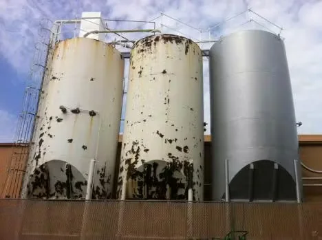 Three large industrial storage tanks showing different stages of corrosion control, with rusted and peeling paint on two tanks and a freshly coated silver tank on the right.