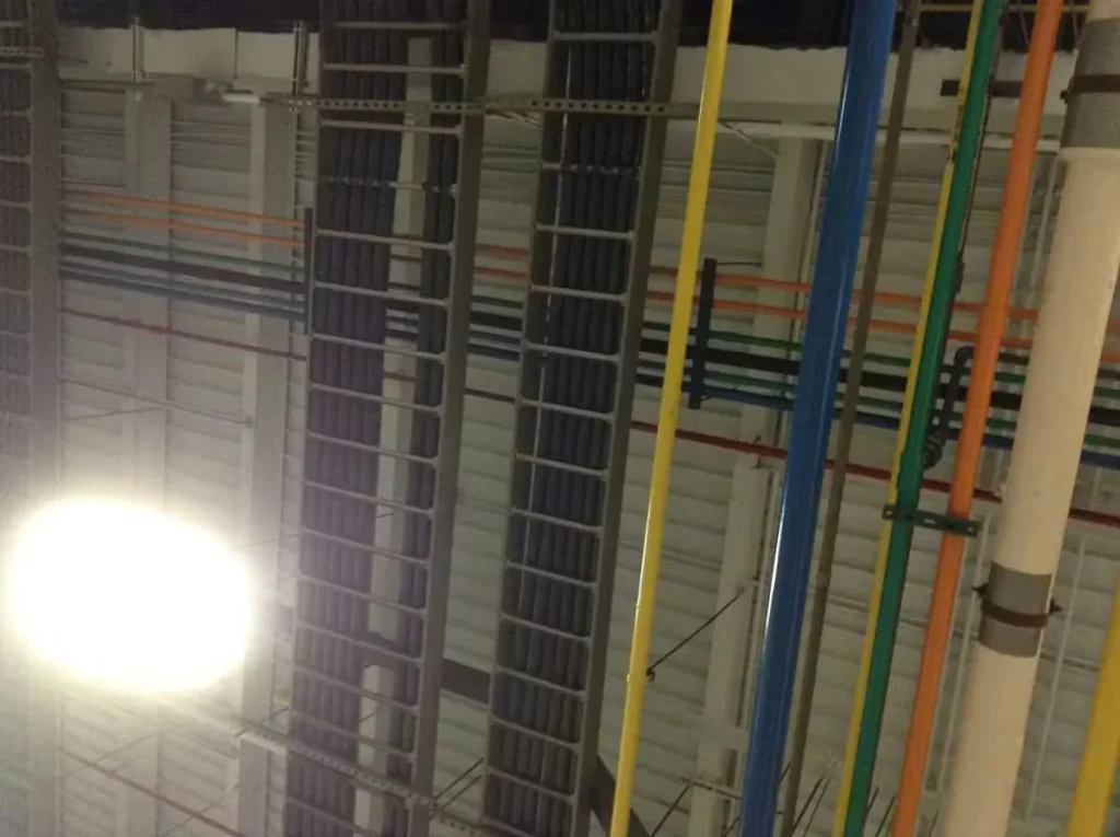 Industrial facility ceiling with exposed steel beams, cable trays, and color-coded pipes showing signs of rust and corrosion near overhead lighting.