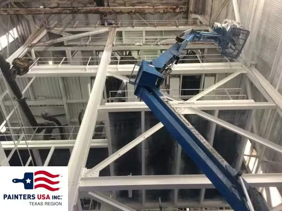 A blue boom lift extends up inside an industrial facility, reaching white steel beams and catwalks near the ceiling; a “Painters USA Inc. Texas Region” logo appears in the bottom-left corner.