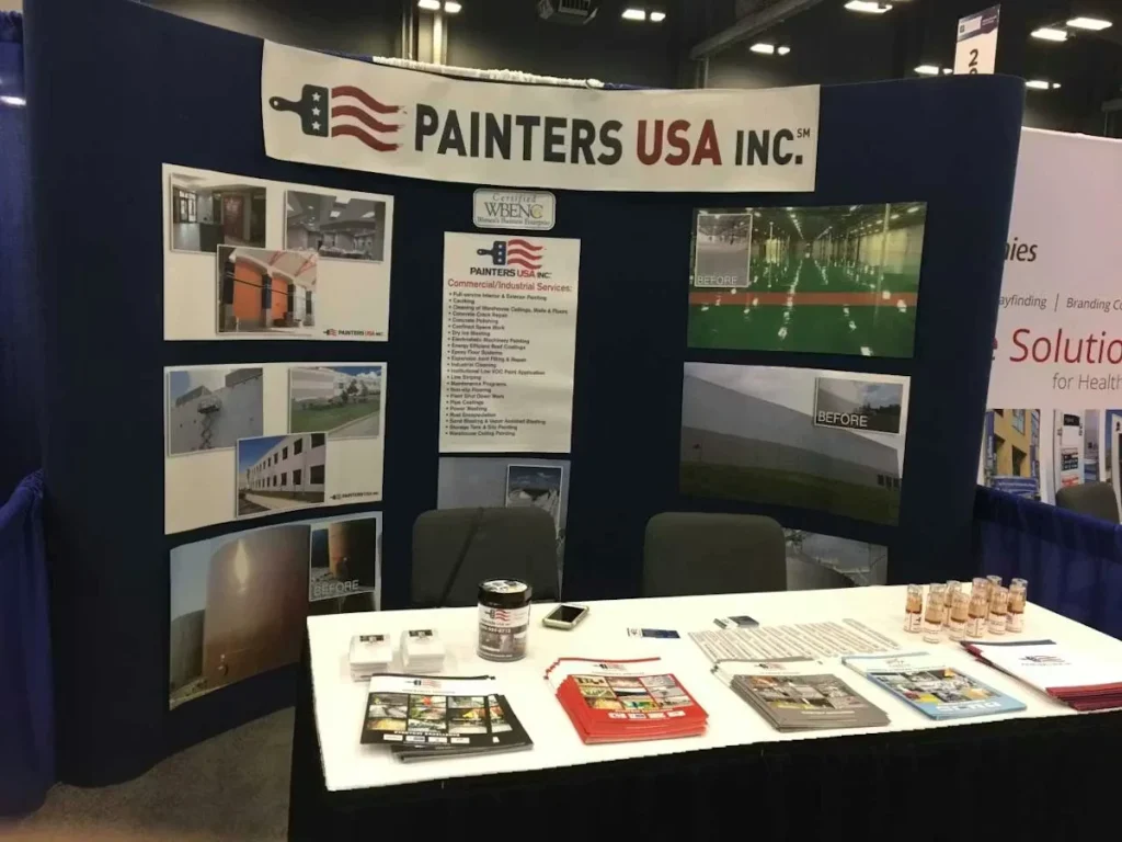 Trade show booth for Painters USA Inc. with a curved backdrop displaying project photos and certifications, plus a table covered with brochures, a phone, and small sample bottles.