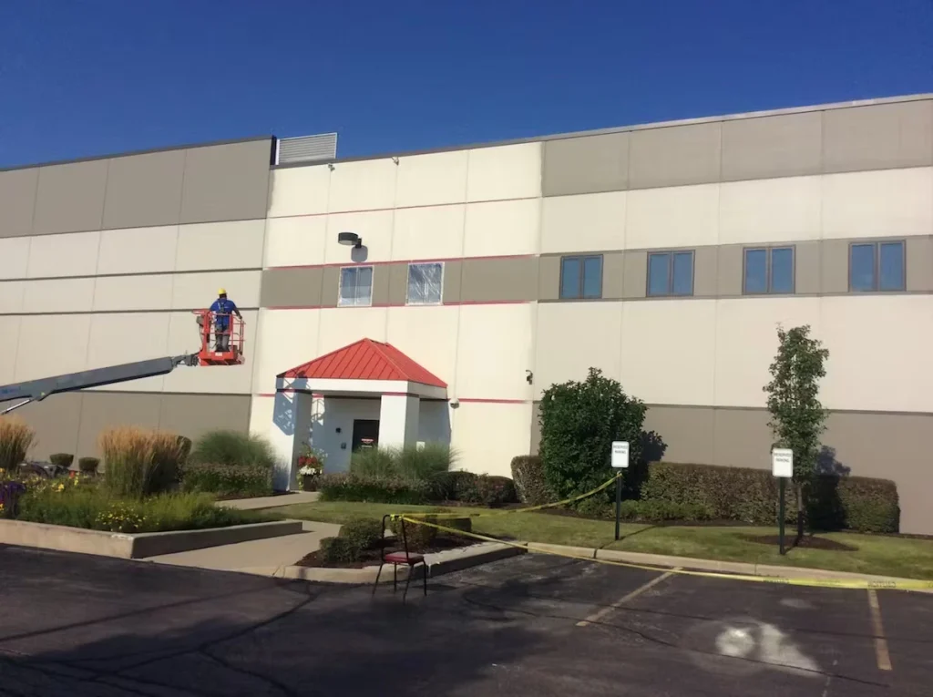 Worker on a boom lift painting or caulking the exterior wall of a large industrial building above a red-roofed entrance, with landscaping and an empty parking lot in the foreground.
