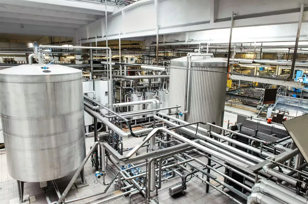 A large brewery production area with stainless-steel tanks, catwalks, and a dense network of pipes and valves spanning the facility.