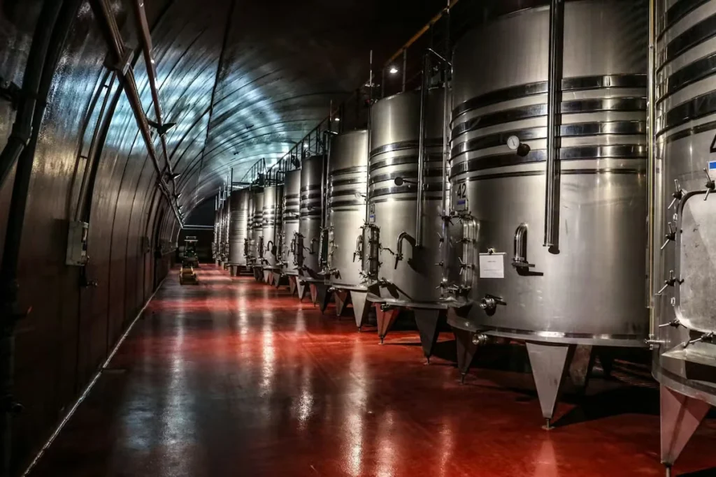 Dimly lit winery or brewery corridor lined with large stainless steel fermentation tanks beside a glossy red floor reflecting overhead lights.