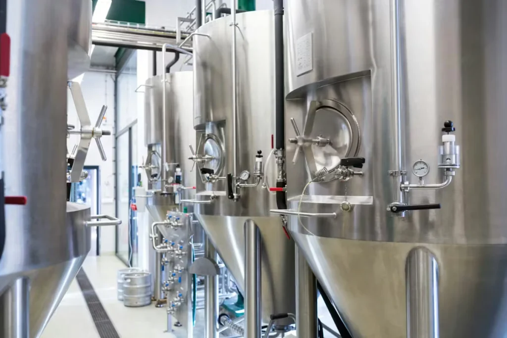 A row of stainless-steel fermentation tanks with valves, gauges, and piping lines a clean brewery production area, with floor drains and a keg visible in the background.