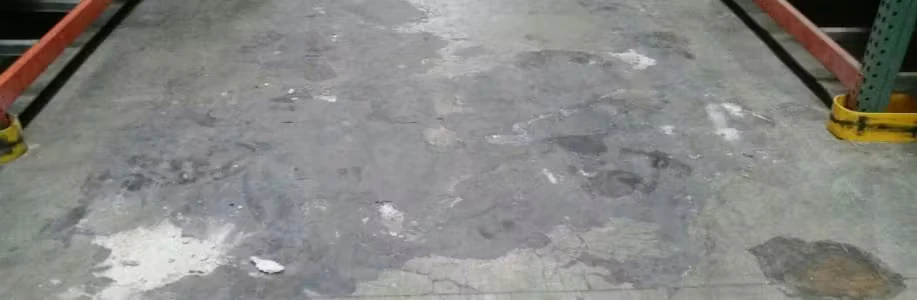 Worn concrete warehouse floor with dark stains, scuffed patches, and visible cracks between two rack bases with yellow guards.