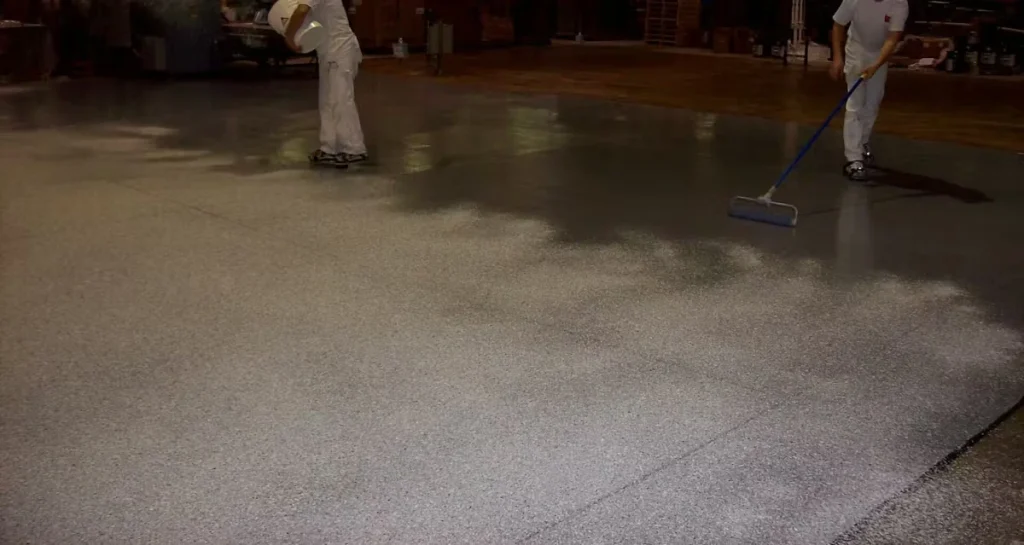 Two workers in white coveralls apply a gray, textured anti-slip coating to a warehouse floor; one pours material while the other uses a long-handled roller/squeegee, with glossy wet sections blending into the rougher finished surface.