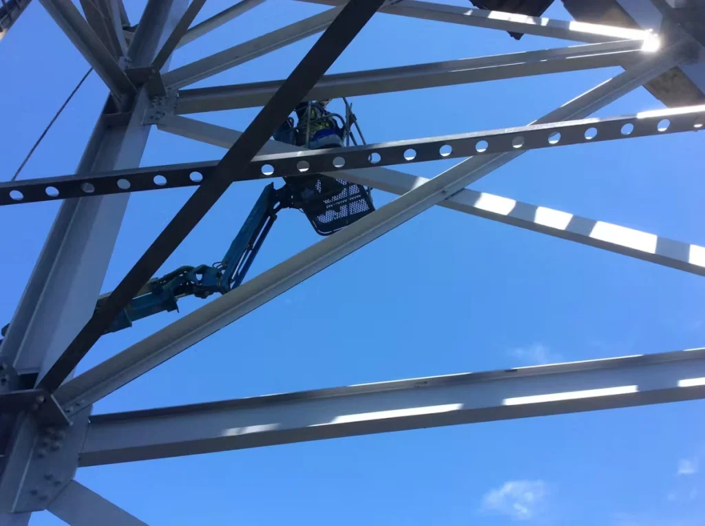 A worker in safety gear stands in a lift basket high above the ground, painting or inspecting steel beams of an industrial structure, with intersecting metal supports framing a bright blue sky.