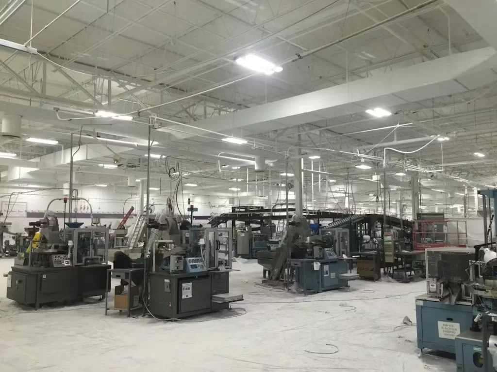 Large industrial factory floor with rows of machinery and conveyor systems under a high white ceiling with bright overhead lights, with dust or debris scattered across the floor.