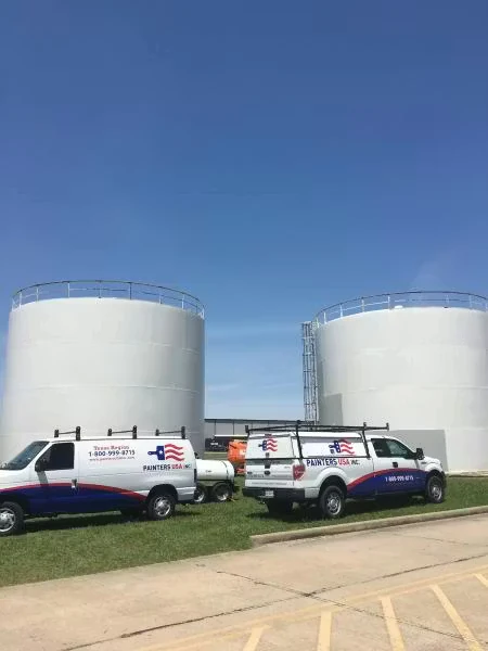 Two white service vehicles with “Painters USA Inc.” branding are parked on grass beside a paved lot, with two large white cylindrical storage tanks and a clear blue sky in the background.