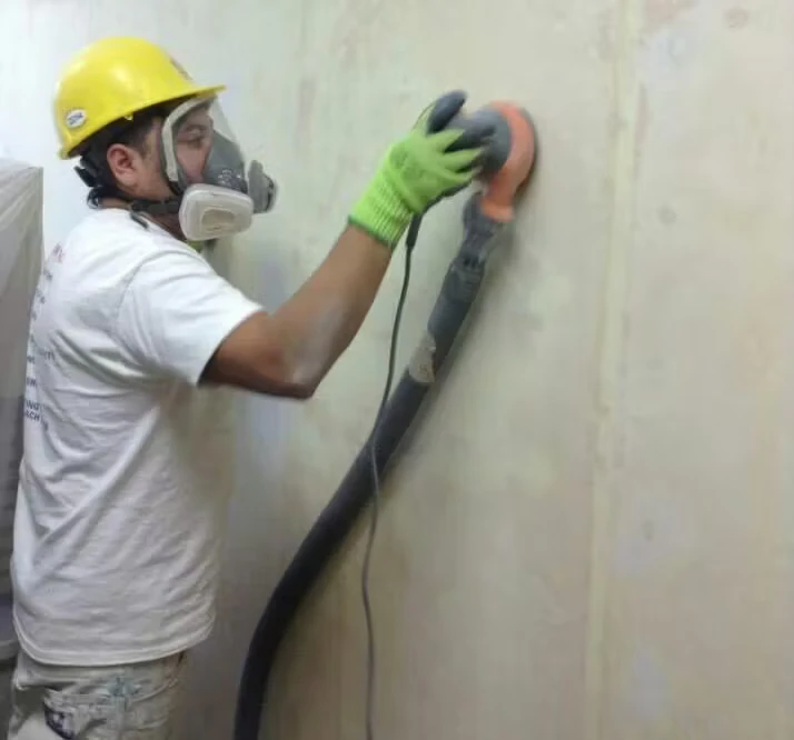 A worker wearing a yellow hard hat, respirator mask, and green gloves sands a wall with a powered hand sander connected to a large vacuum hose.