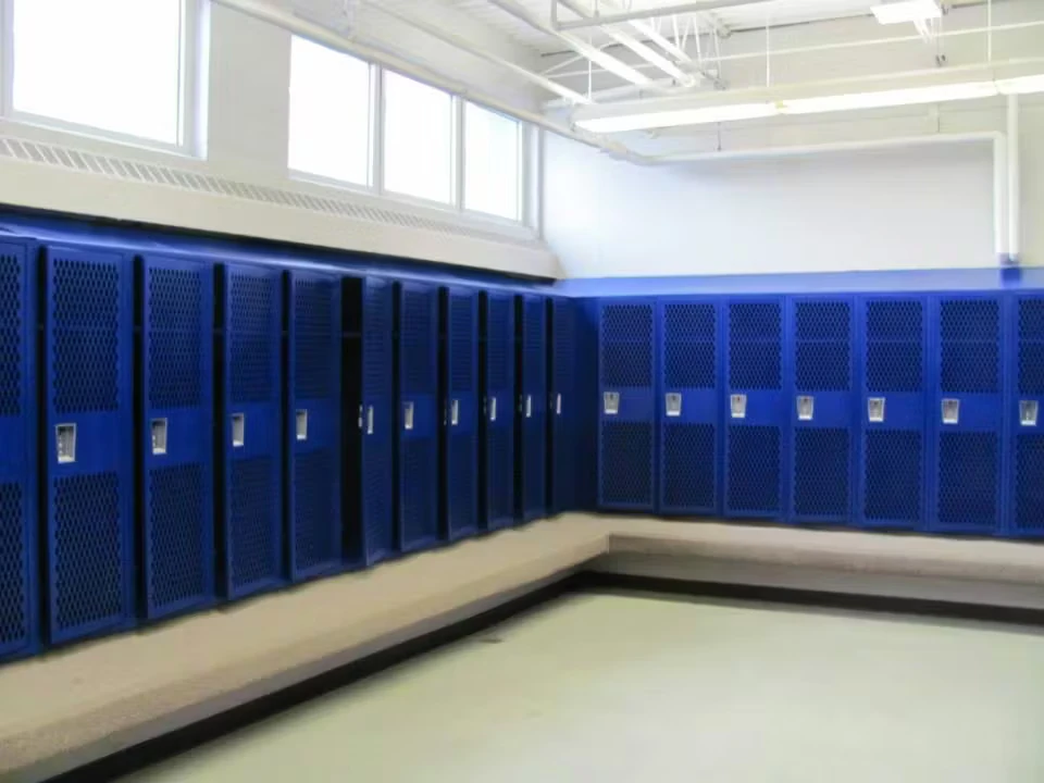 A bright locker room with rows of blue metal lockers along two walls, high windows letting in daylight, and a long built-in bench wrapping around the corner above a light-colored floor.
