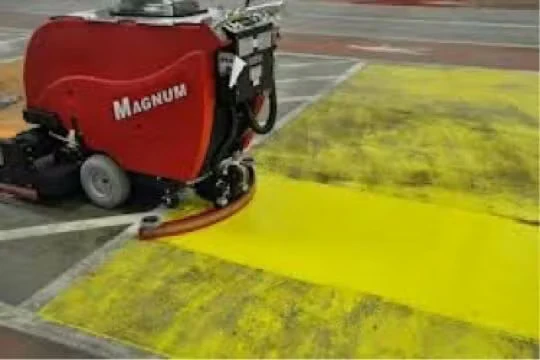 A red industrial floor scrubber cleaning a concrete floor, leaving a bright yellow cleaned stripe across a darker, dirtier surface.